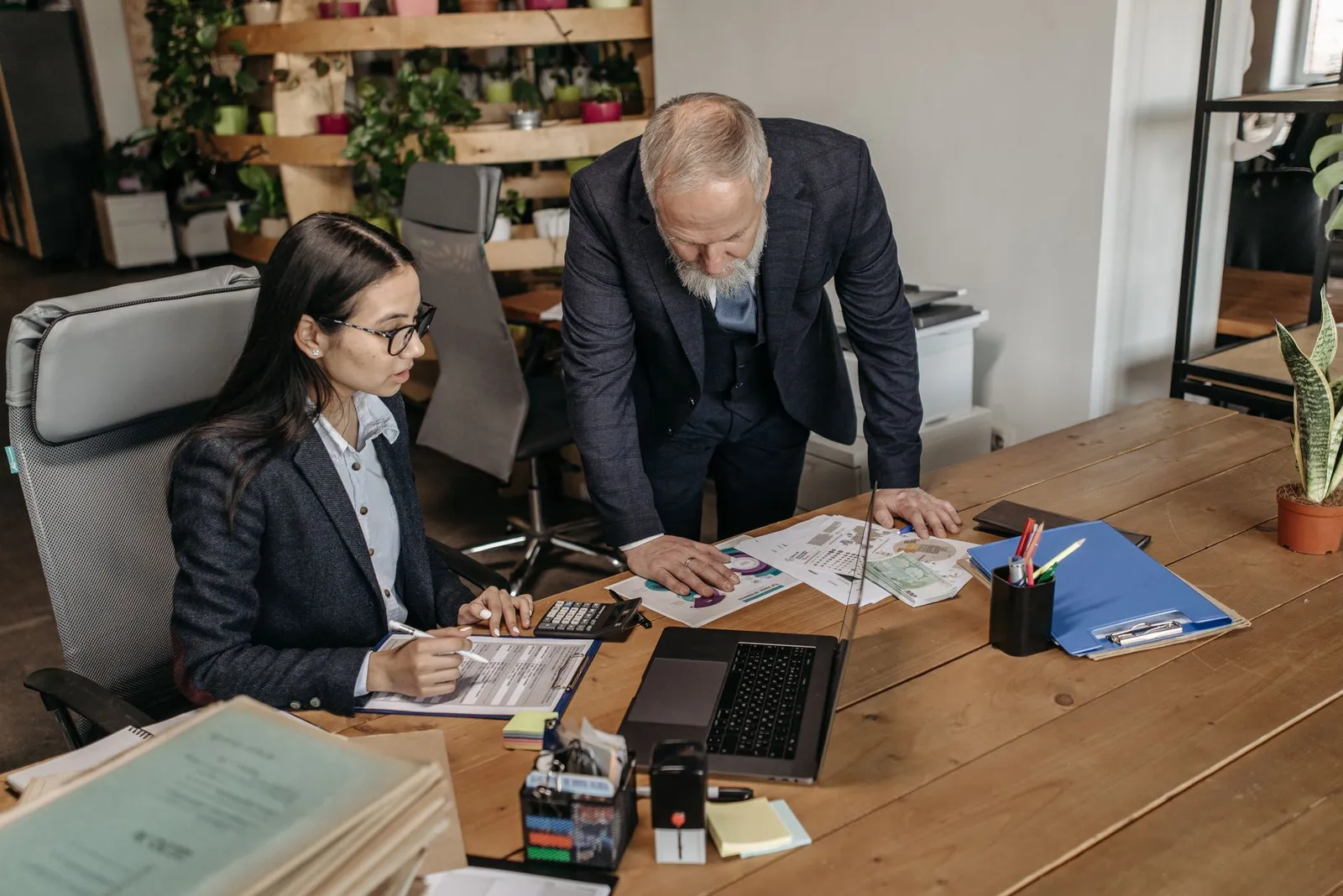 Two colleagues at a wooden desk going through printed documents beside a laptop and calculator.