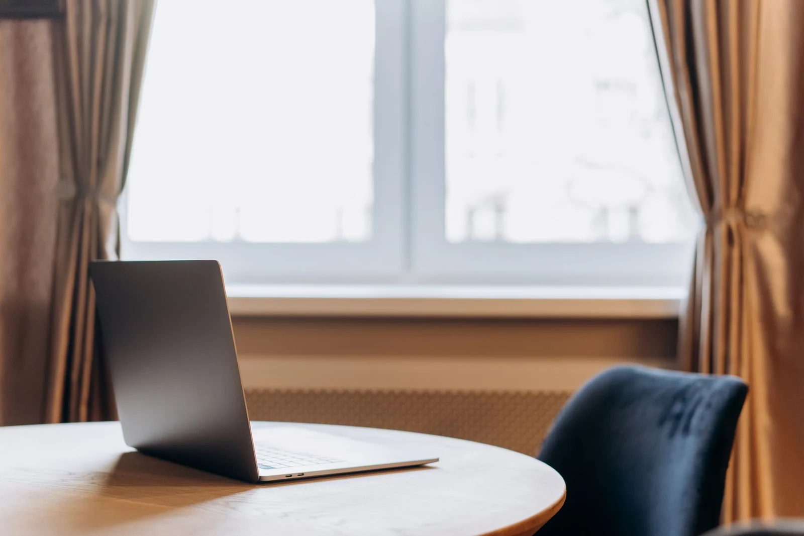 An open silver laptop on a round wooden table beside a curtained window, with an empty blue chair pulled up to it.