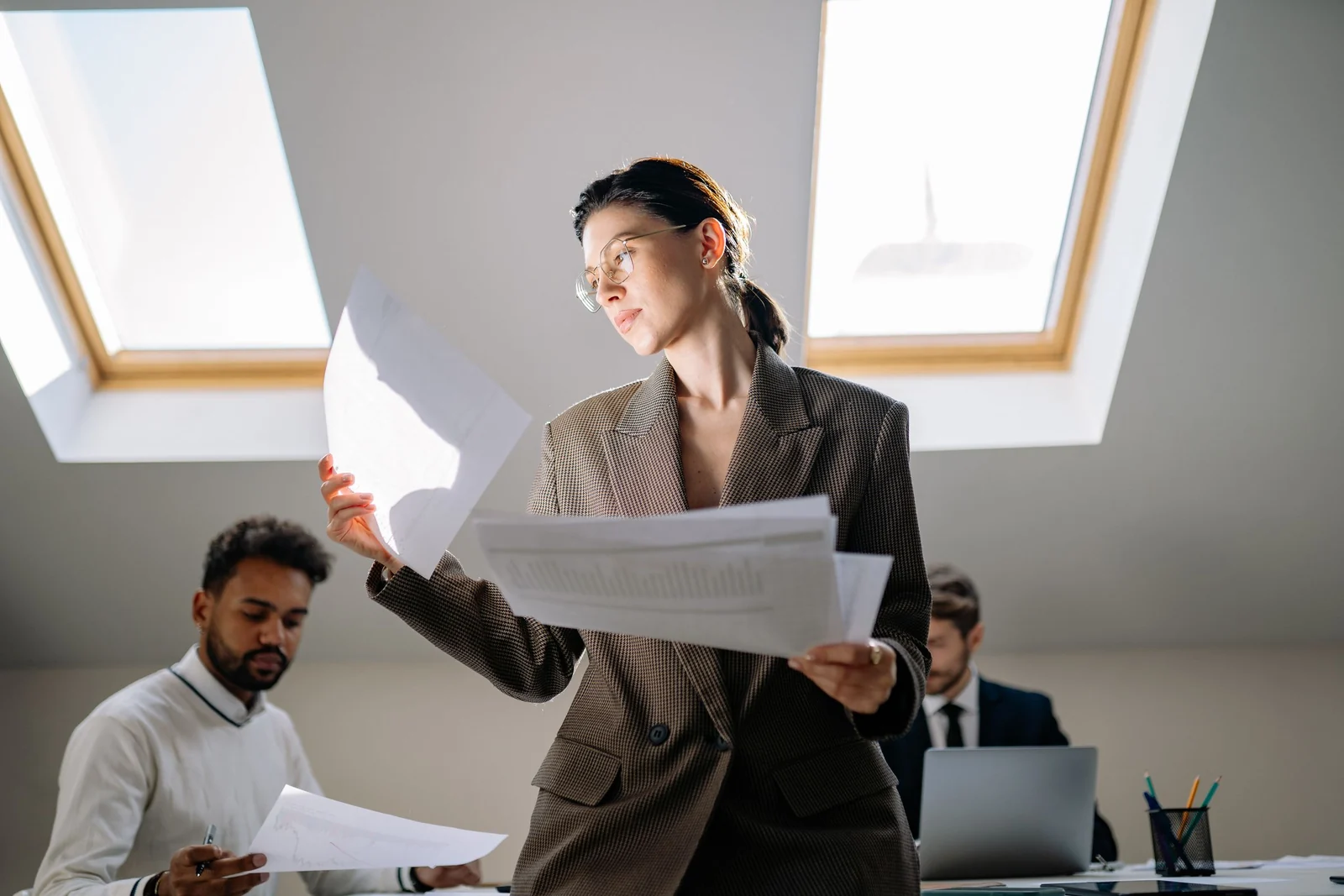 Woman in an office holding up two printed sheets and comparing them, colleagues working behind her