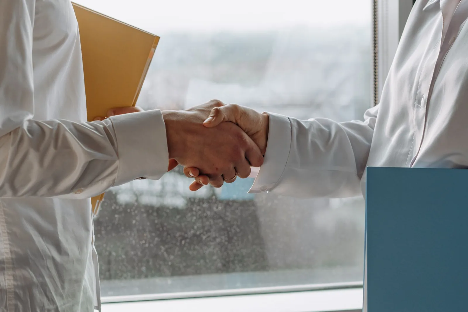 Two people in white shirts shaking hands beside document folders at an office window