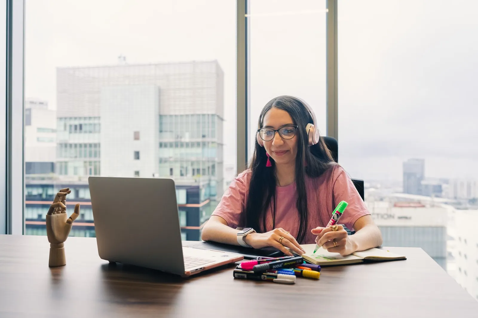 Woman in a high-rise office working at a laptop, with the city visible through the window behind her.