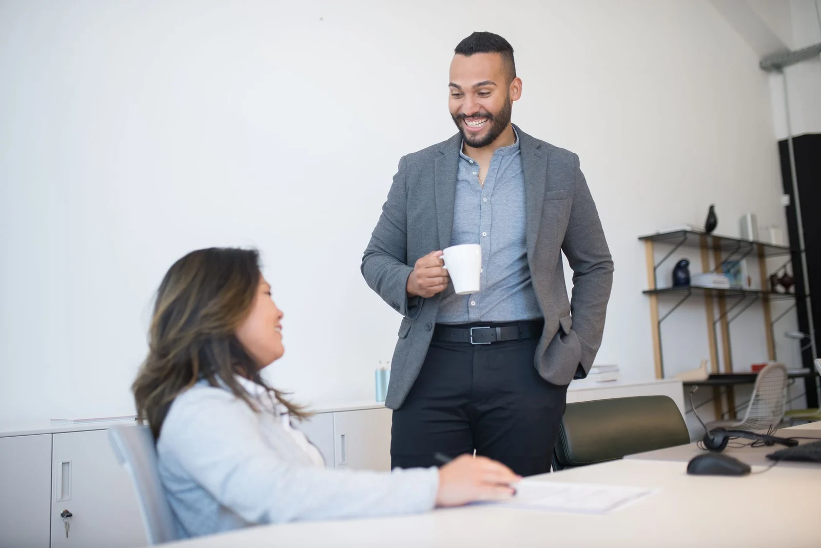 A man holding a mug talking with a seated colleague who is writing on a document in a bright white office