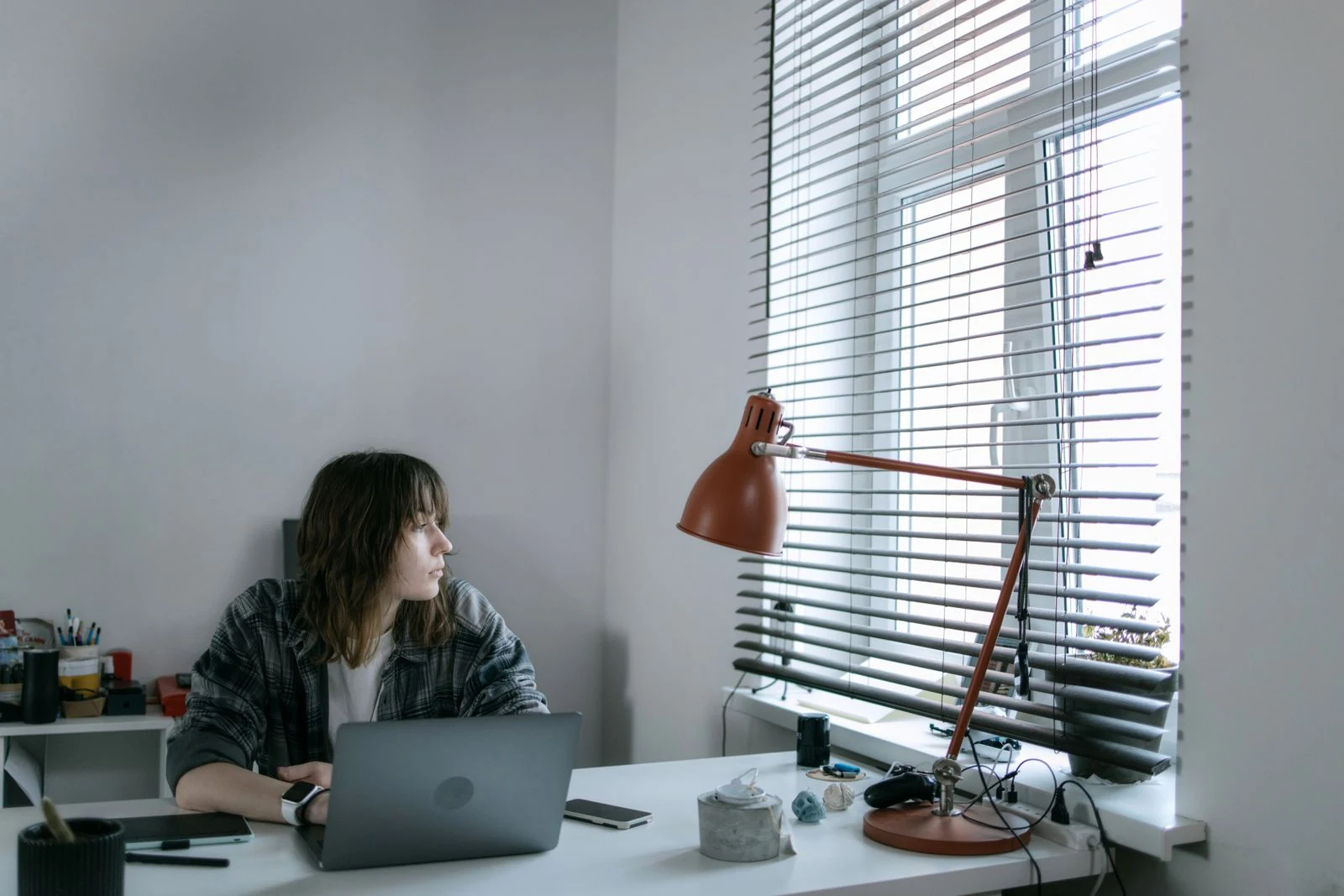 A woman sitting at a desk with a laptop, turned away from the screen and looking out through window blinds.