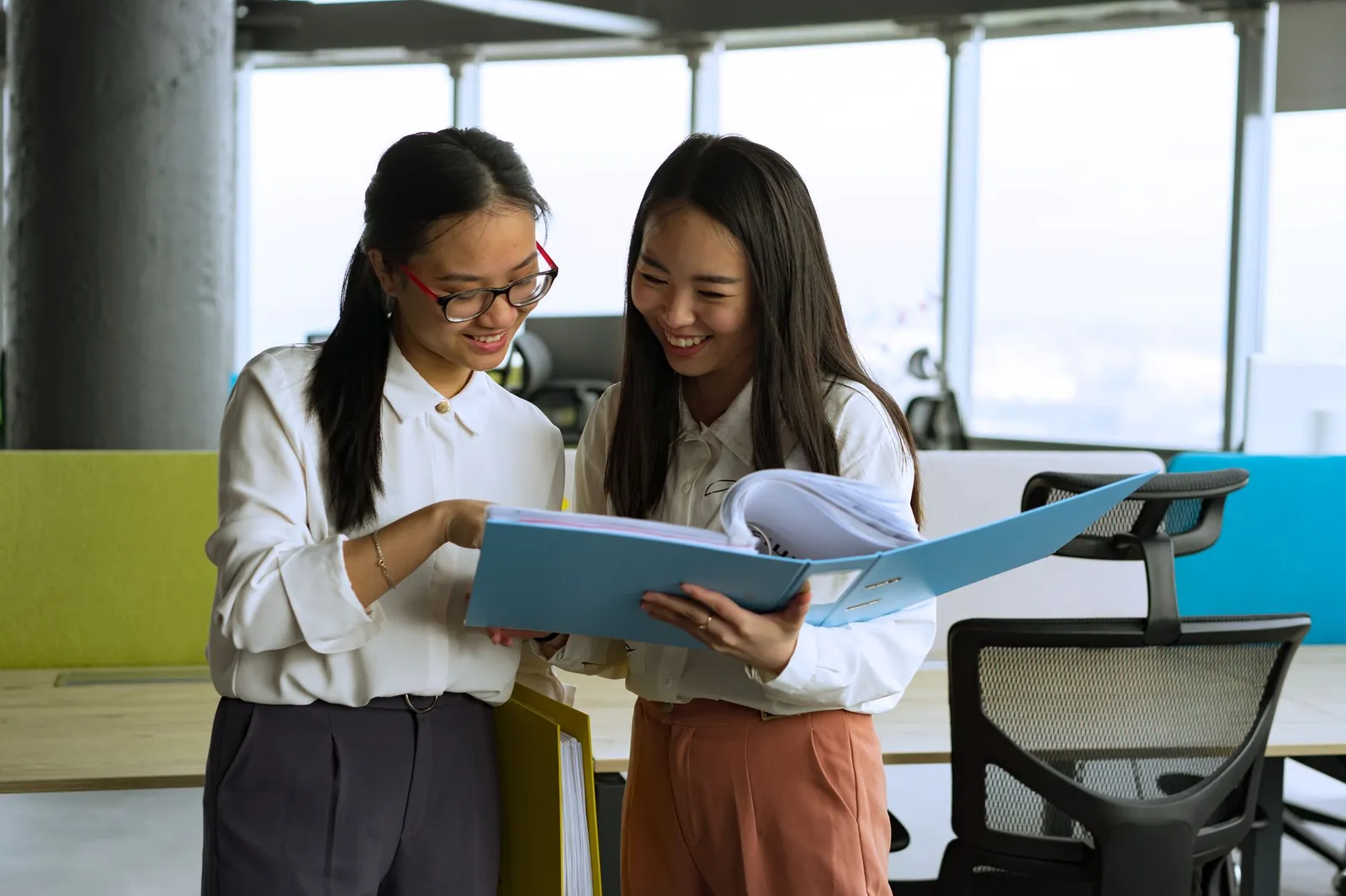 Two colleagues standing in a bright open-plan office, reading through a blue folder of printed documents together