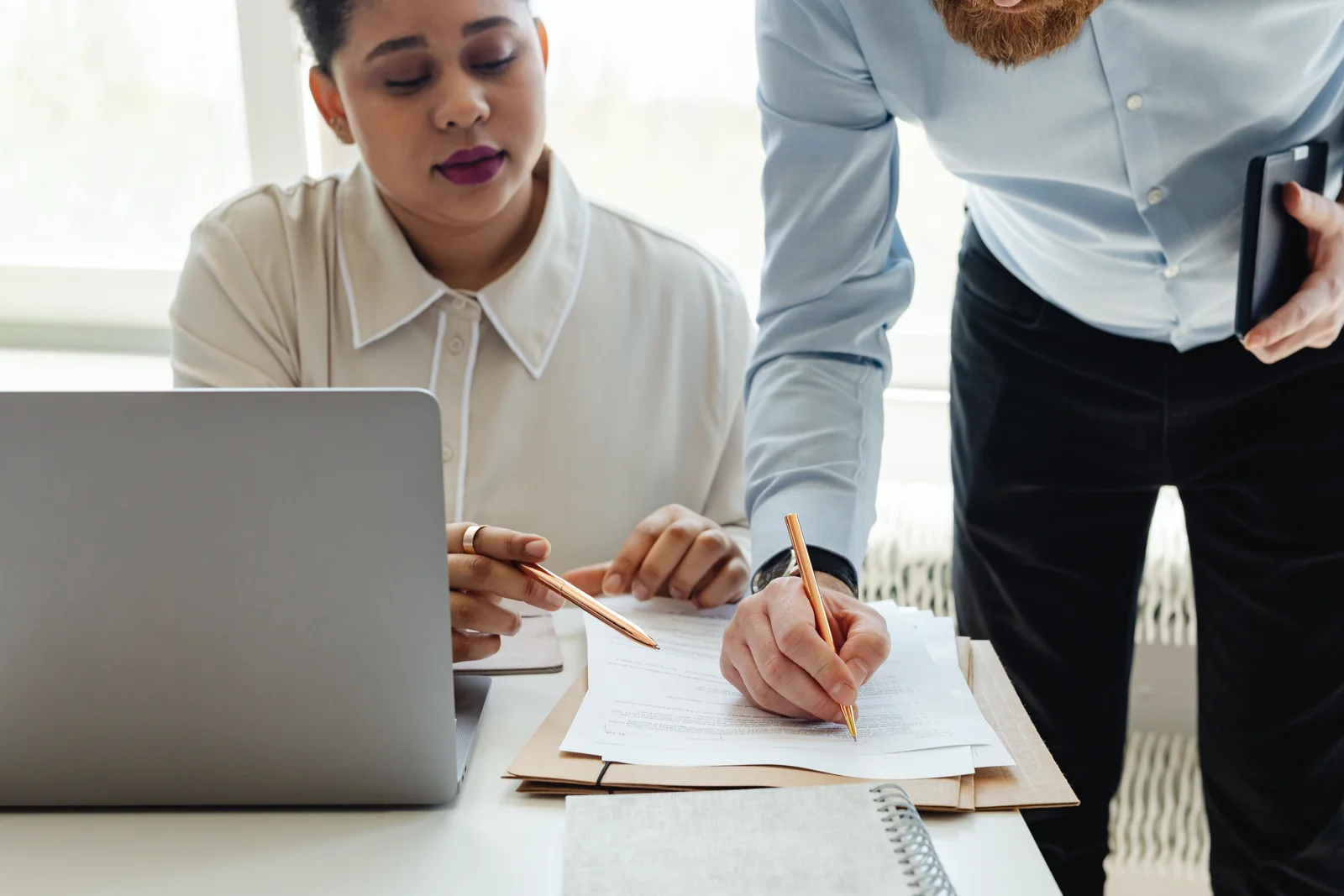 Two colleagues at a bright office desk working through printed documents with a laptop open beside them