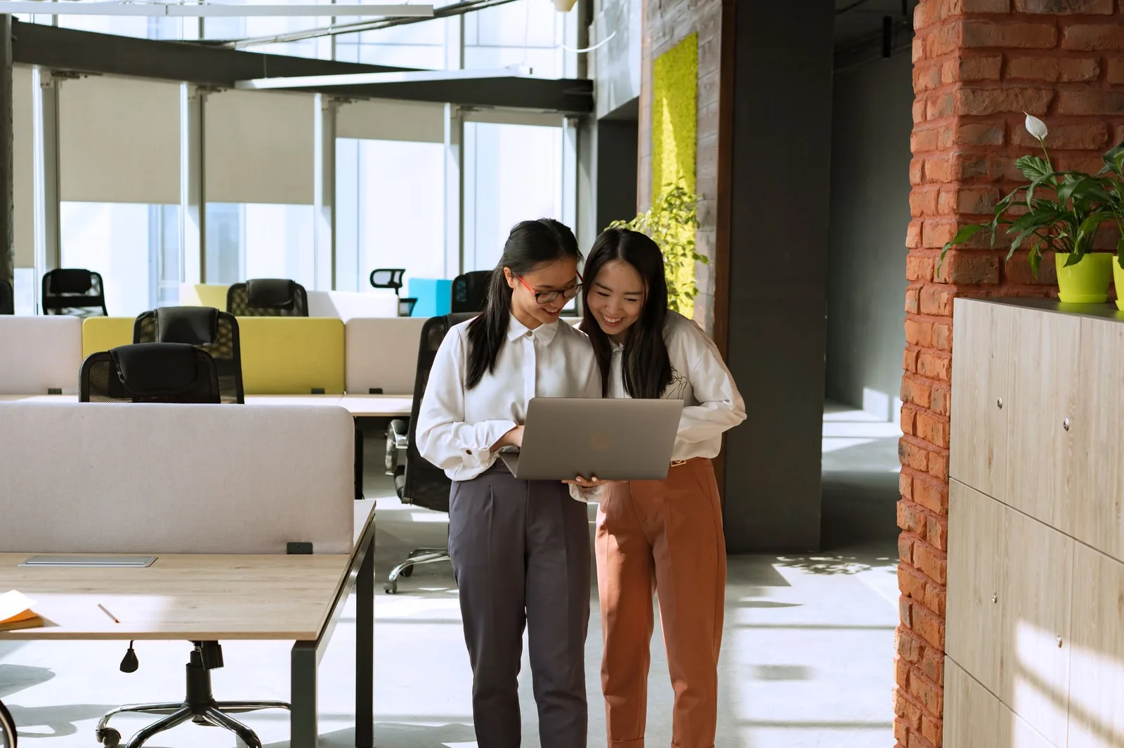 Two colleagues standing with a laptop in an open-plan office, a bank of empty desks and chairs behind them