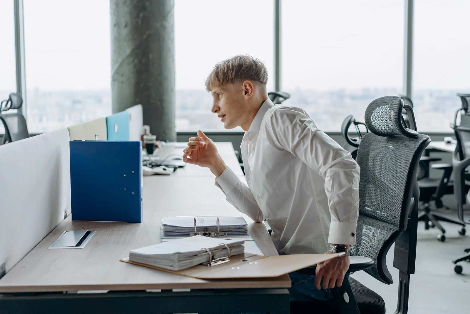 Man at an open plan office desk leaning over stacked ring binders and loose paperwork