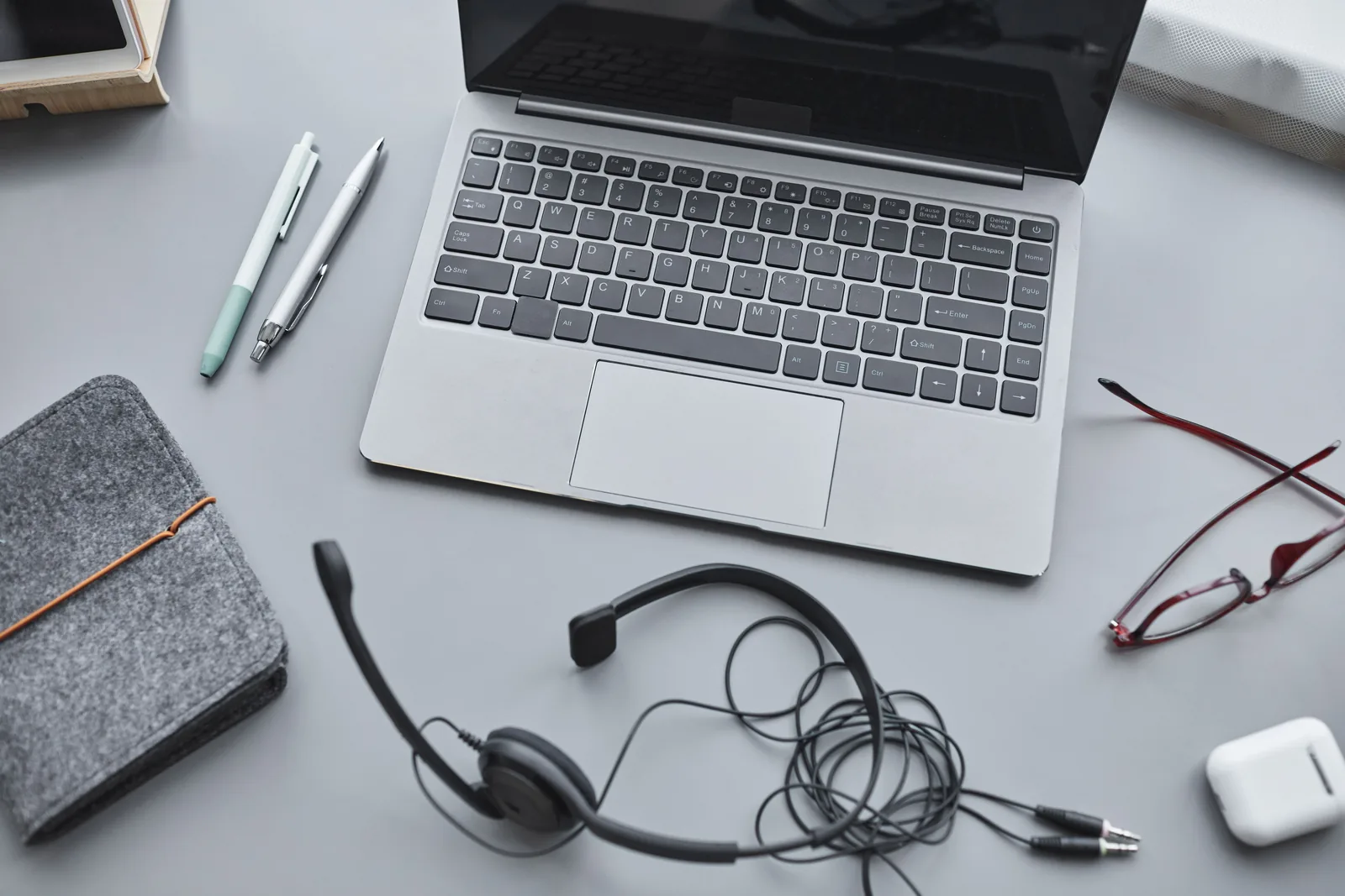 Overhead view of a desk with an open laptop, a wired headset, earbuds, reading glasses, two pens and a felt notebook sleeve