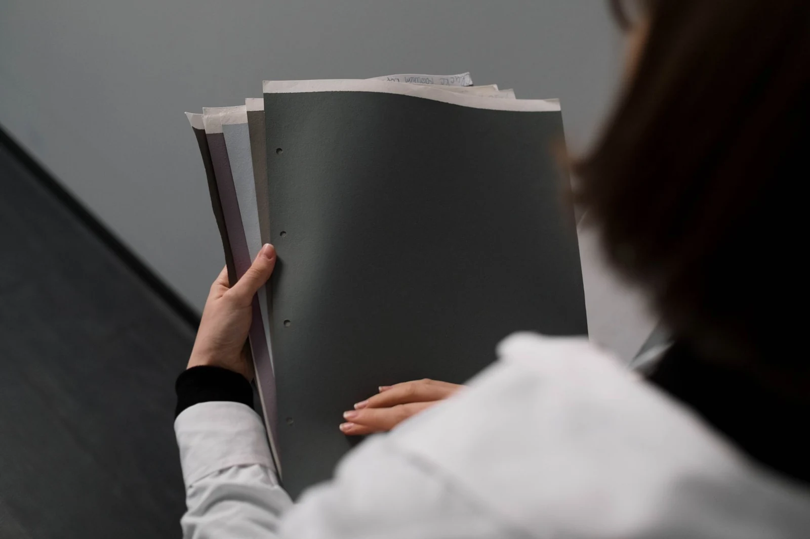 Over the shoulder of a woman in a white shirt holding a stack of document folders against a grey office wall