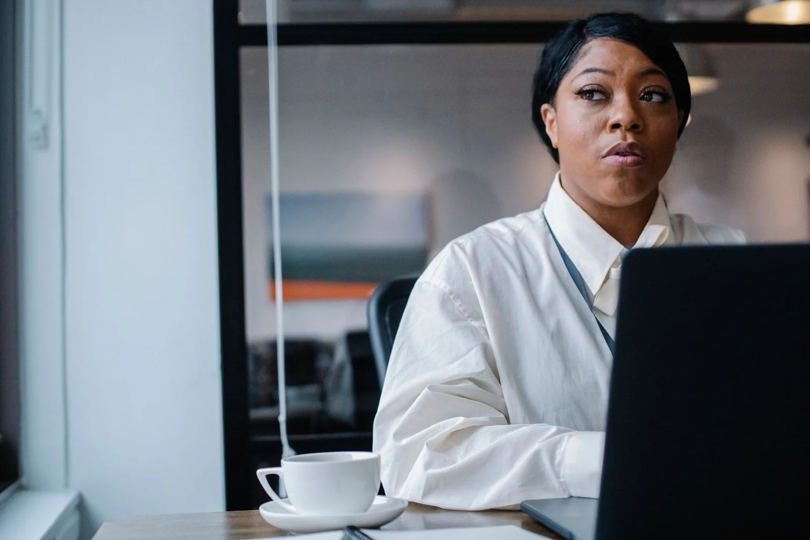 Woman at an office desk beside a laptop and a coffee cup, looking away in thought