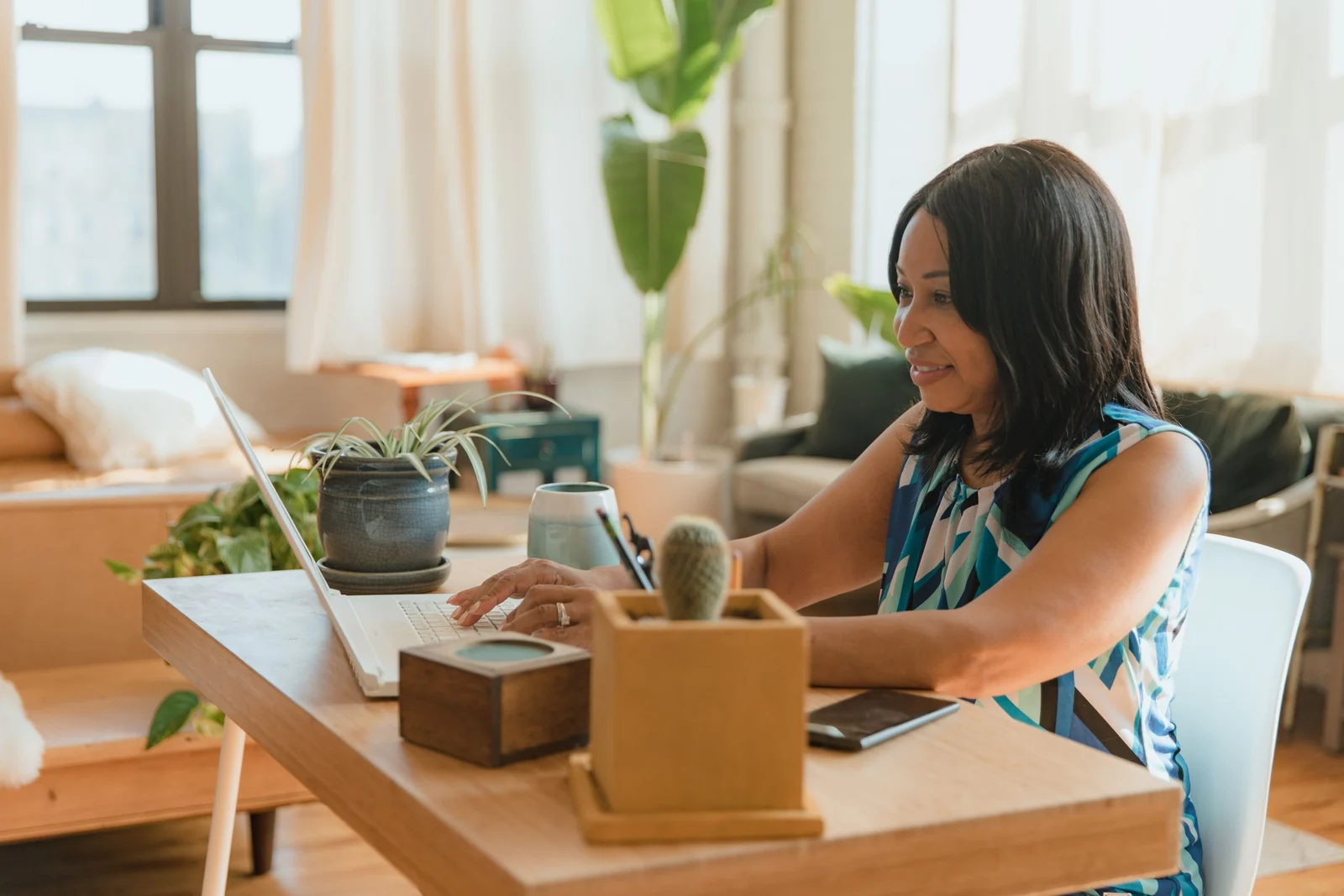 Woman working on a laptop at a wooden table in a bright living room with plants