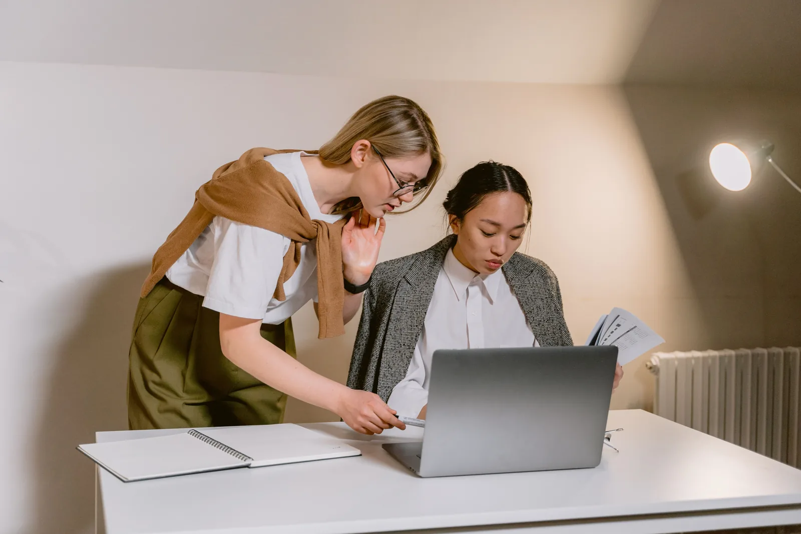 Two colleagues at a white desk in an office, one holding a printed report while the other works on a laptop