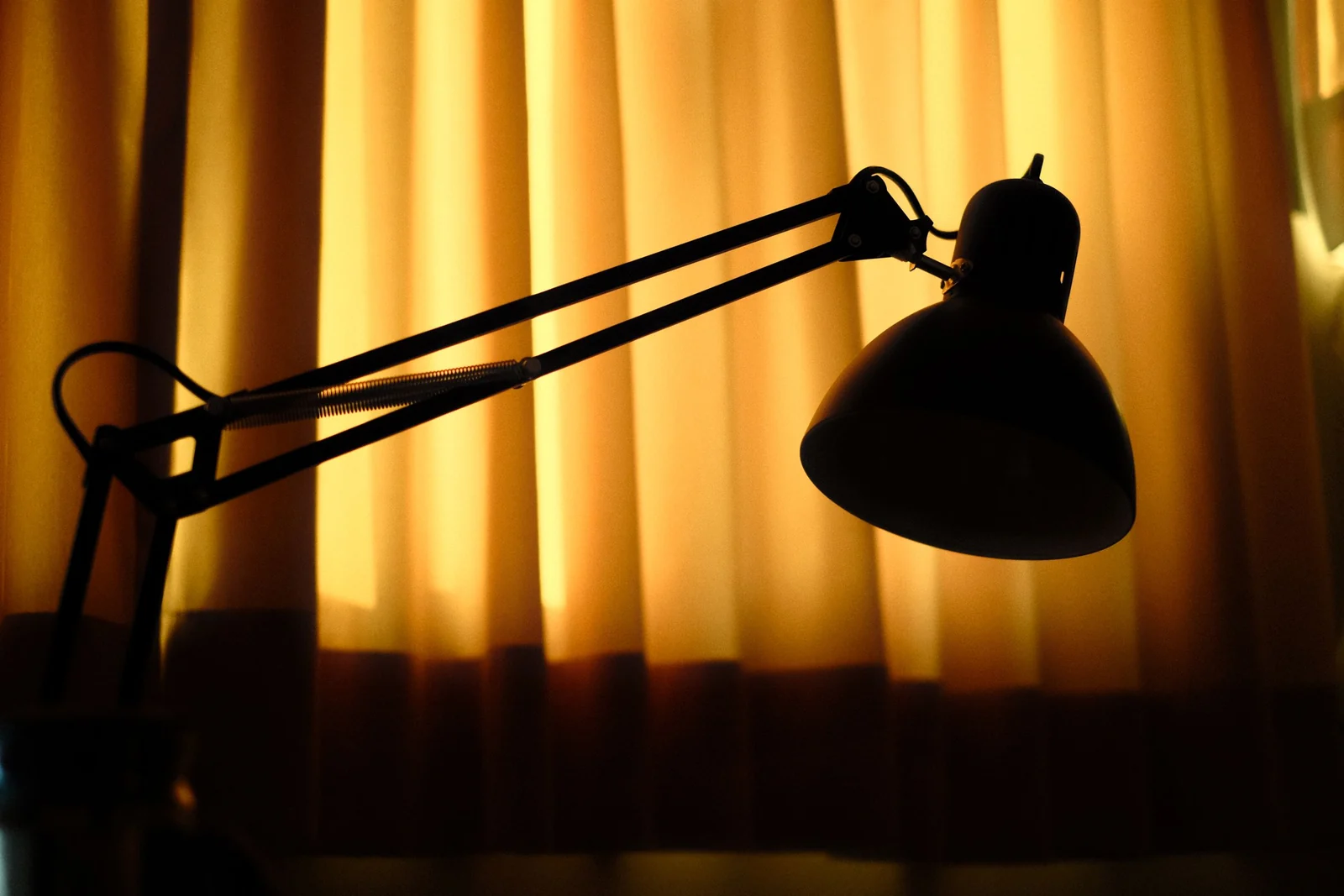 Angled desk lamp silhouetted against warm backlit curtains in a dim home workspace